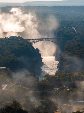 Aerial view shows Victoria Falls Bridge on the Zimbabwe Zambia border at sunset, spray rising from Victoria Falls, steel arch silhouetted, river glinting, rainforest and village roofs visible.