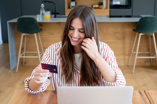 Smiling woman using a credit card and laptop for online banking at home kitchen table
