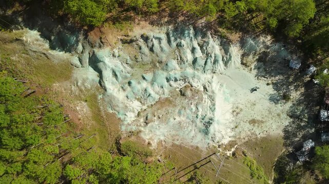 Aerial top-down rise above Blue Soil Hills in Sagada reveals full extent of surreal blue-green mineral clay formation surrounded by pine forest with tourists exploring on sunny day
