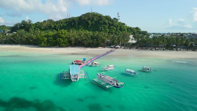Aerial orbit around colorful long pier extending into turquoise waters of White Beach Boracay, with multiple bangka outrigger boats moored alongside and lush hill behind