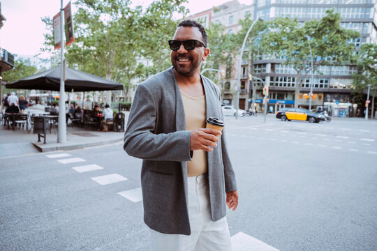 Smiling latin american man crossing street with disposable coffee cup, expressing urban lifestyle and happiness.