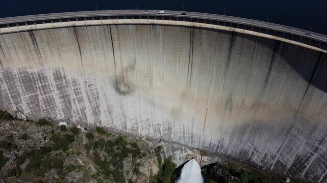Spectacular 4K drone shot advancing and descending over the massive concrete wall of the Almendra Dam in Salamanca, Spain. Featuring cars on the crest road and a high-pressure water release below