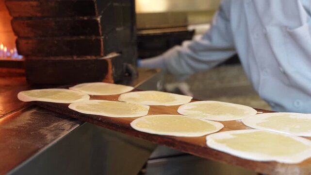 A gloved chef prepares traditional flatbread in a commercial kitchen. Close-up cinematic shot shows brushing dough with oil and sprinkling seeds on a wooden paddle before baking in a wood-fired oven.