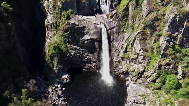 4K cinematic aerial drone shot slowly approaching the spectacular Pozo de los Humos waterfall in Salamanca, Spain. High-pressure water bursts through the rocky cliff and plunges into the dark lagoon b