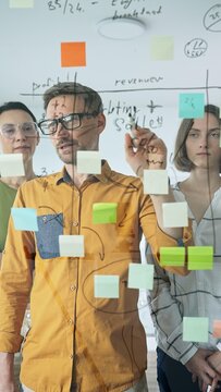 Business people team brainstorming, sharing strategy on glass whiteboard with colorful sticky notes, working collaboratively in bright office