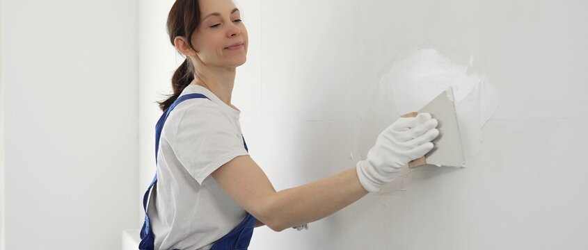 Construction worker woman spreading finishing putty on white wall, wearing protective gloves while smoothing surface with putty knife for precise home repair