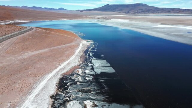 Panoramic pan over Laguna Tara deep blue waters with jagged salt crust shoreline white flats red desert terrain and distant Andean peaks under clear sky. Laguna Tara, San Pedro de Atacama, Chile