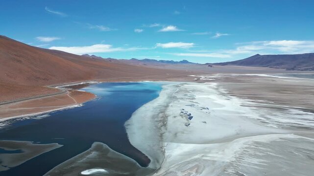 Panoramic aerial shot of Laguna Tara deep blue waters beside vast white salt flat with red volcanic hillside and Andean mountain range under blue sky. Laguna Tara, San Pedro de Atacama, Chile
