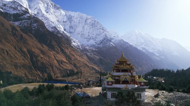 Breathtaking aerial drone view of a Tibetan Buddhist monastery in Nepal's Manaslu region, dwarfed by towering snow-covered mountain walls rising dramatically into a hazy blue Himalayan sky.