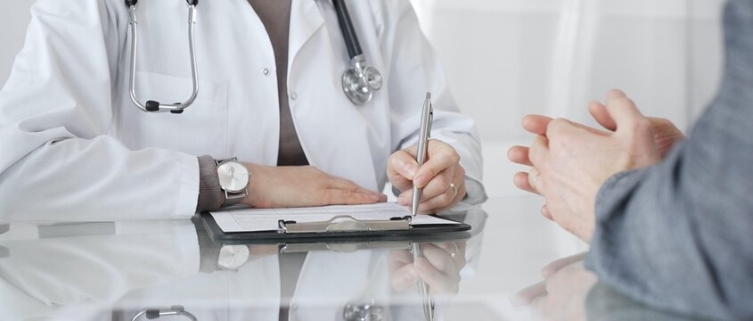 Doctor consulting patient, physician writing medical notes during healthcare examination in modern clinic office. Medicine and health care