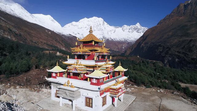 Aerial drone footage of an ornate red and gold Tibetan Buddhist monastery nestled in Nepal's Manaslu valley, with the majestic snow-capped Manaslu mountain range rising dramatically in the background 