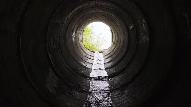 Water flowing along a textured concrete drain pipe towards the bright opening, with green leaves visible at the tunnel's end, creating a striking contrast between darkness and escape