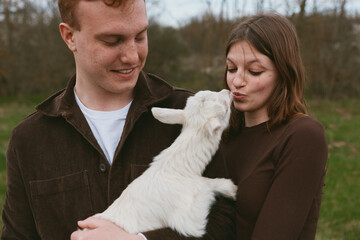 Fototapeta premium Candid lifestyle shot of a couple with a playful goat kid outdoors, the pair share affection as the animal nuzzles them, rustic field setting, relaxed weekend moment.