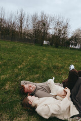 Fototapeta premium Lifestyle scene shows a couple lying on grass outdoors with a curious goat kid, wearing cozy knitwear, sharing affectionate smiles in a relaxed countryside moment under soft daylight.
