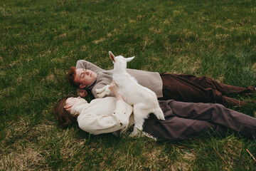 Fototapeta premium Relaxed couple lying on grass with a curious goat kid climbing nearby. Warm earthy tones highlight outdoor lifestyle, companionship, and gentle play. Soft daylight creates a cozy, candid moment of