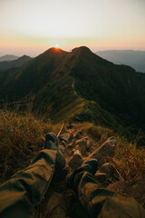 Fototapeta premium The frame shows the hiker's legs in green trekking pants and sturdy hiking boots resting on rugged rocks and dry grass in the foreground. A narrow,
