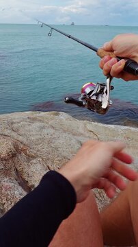 Vertical video. First Person View of Fisherman Casting Spinning Rod From Rocky Tropical Coast Into Calm Ocean Water Under Cloudy Sky, Active Saltwater Fishing Adventure