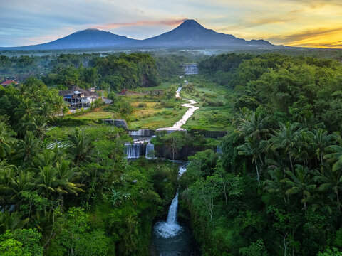 Aerial view of Grojogan Watu Purbo multi-tiered waterfall surrounded by lush tropical greenery with Mount Merapi in the background during sunrise Kecamatan Tempel, Daerah Istimewa Yogya, Indonesia.