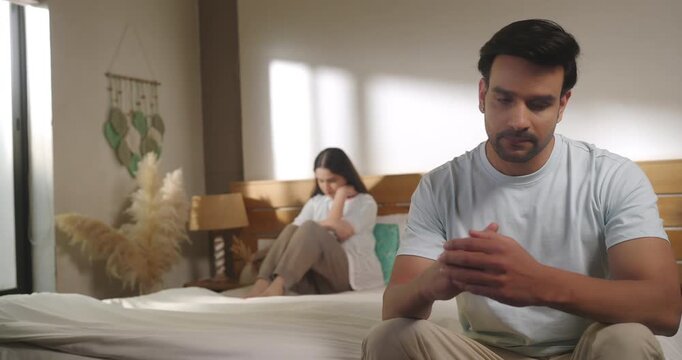Indian young adult gen z upset male female couple pair quiet sitting on bed feel bad hurt think deep have family issue tense tired man woman look sad show distance after fight argue lost job room home
