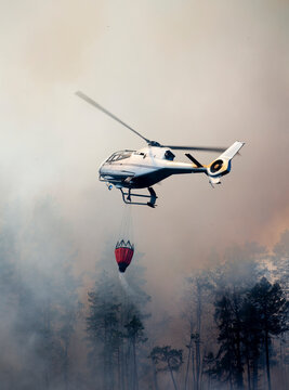 Drone view of helicopter pouring water with bucket on forest fire while flying in smoky sky