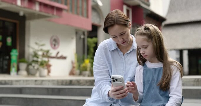 Portrait of mother shows photo on smartphone to little daughter as sits on parapet. Woman shares online content with lovely girl on city street. Digital device
