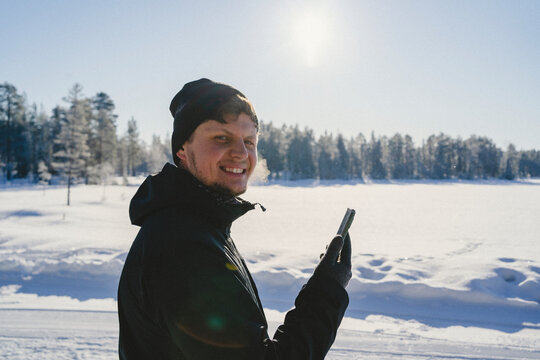 Side view of smiling boy in warm clothes holding smart phone while standing on snow