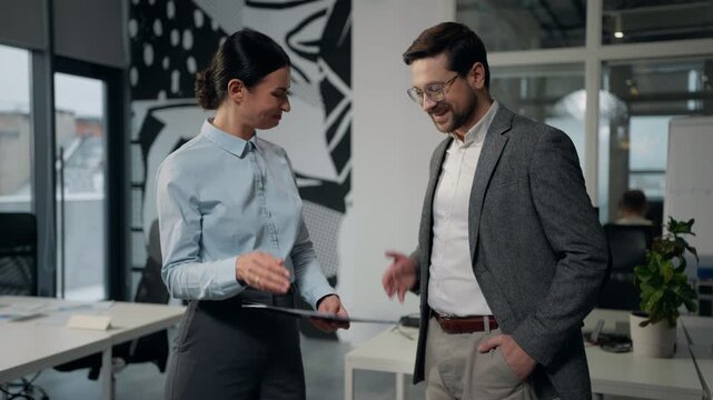 Two Caucasian business colleagues talking looking in folder with papers handshaking leaving office Caucasian businesswoman reading document shaking hands with European businessman agreement going away