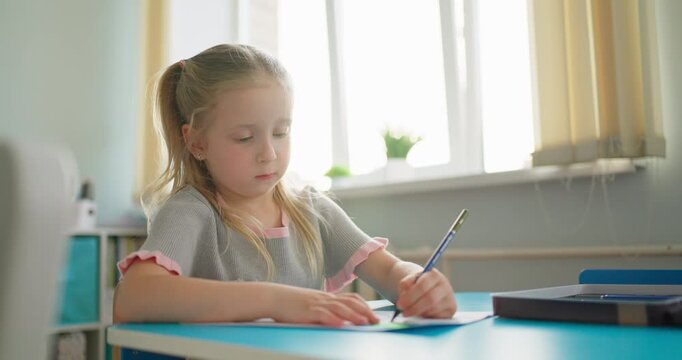 Young schoolgirl concentrating on writing with a pencil at a classroom desk, illuminated by soft daylight from large windows during a quiet lesson