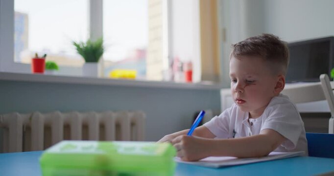 Young schoolboy concentrating on writing in a notebook at a classroom desk, with soft daylight from a window and a pencil case nearby