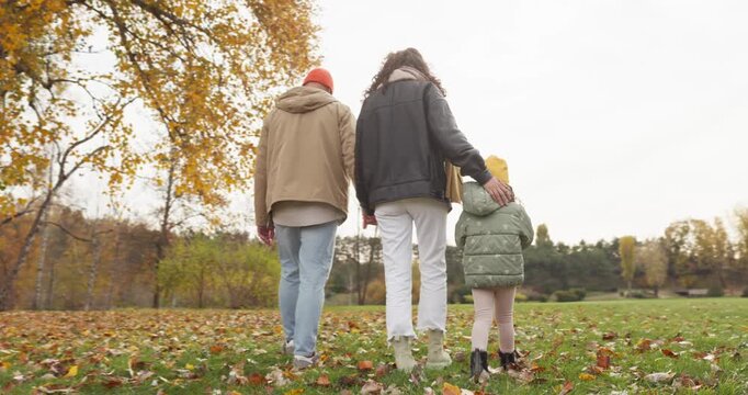 Family with parents and child in autumn park. They walk on colorful leaves in warm clothing, enjoying nature and quality time across a quiet meadow. Concept of love, care, unity and wellness.