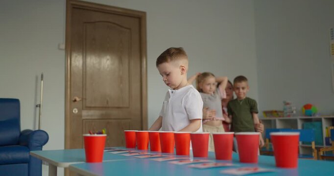 Children line up in a colorful playroom for a red cup challenge, with a focused young boy standing beside a table during an indoor party game