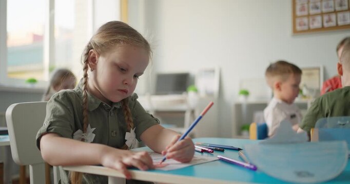 Young girl with braided hair concentrates on drawing with colored pencils at a classroom desk while other children learn in soft background light