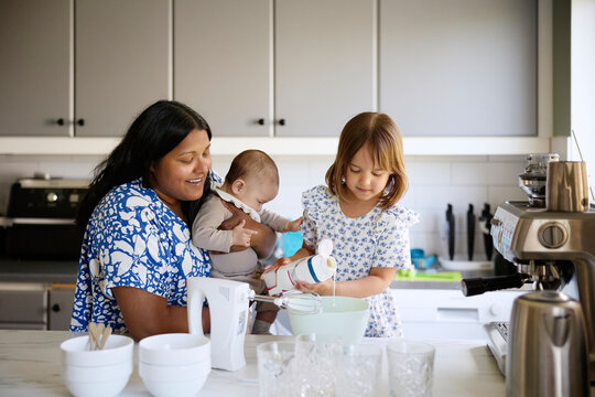 Mother carrying toddler son while assisting daughter pouring cream in bowl at kitchen island