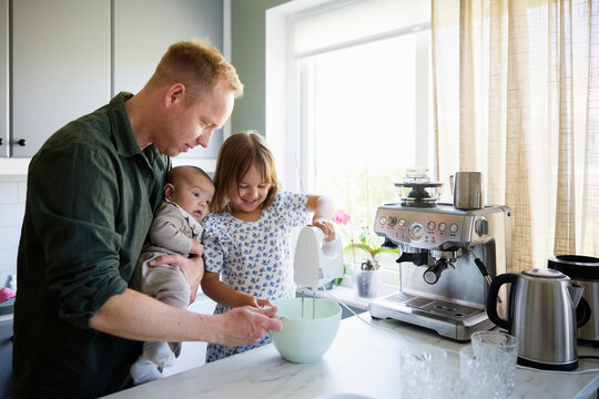 Happy girl using electric beater with assistance of father standing beside with toddler in kitchen