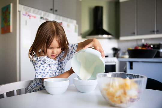 Cute girl pouring cream in bowl while sitting at table in kitchen