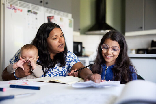 Mother sitting with male toddler while helping daughter in studies at home