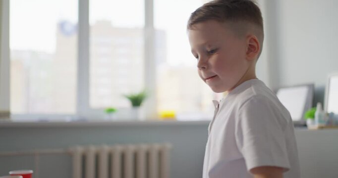 Young boy in white shirt standing at a table with a red cup in a bright room, concentrating during a simple indoor activity near large windows