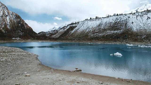 Drone footage of a serene blue glacial lake at the foot of a snow-dusted ridge in Nepal's Manaslu region, with floating chunks of ice along the shore.