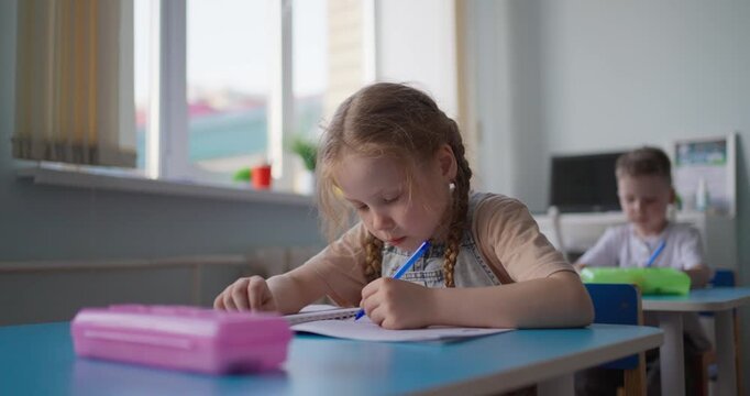 Young schoolgirl with braided hair concentrating on writing in a notebook at a blue classroom desk beside a pink pencil case and sunlit window