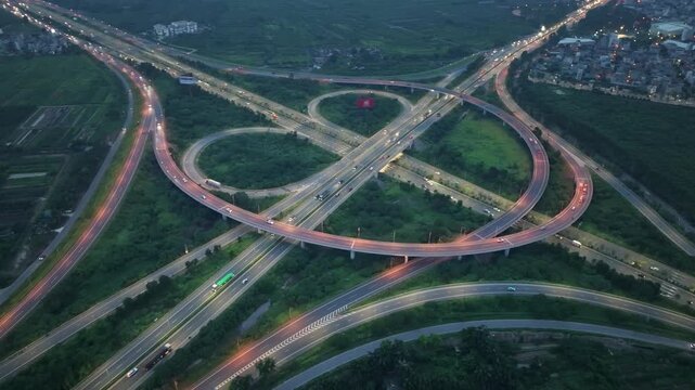 Blue hour drone scene of Co Linh interchange with glowing vehicle light trails crossing multilane expressways and suburban Hanoi residential expansion.