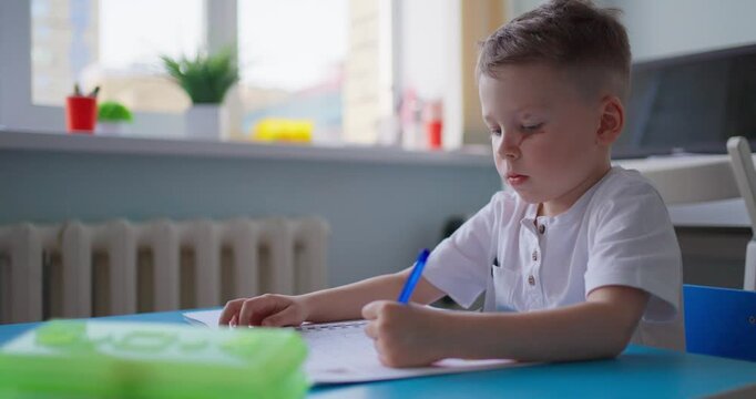 Young boy concentrating on writing in a notebook at a blue classroom desk, practicing schoolwork in a bright learning room by the window