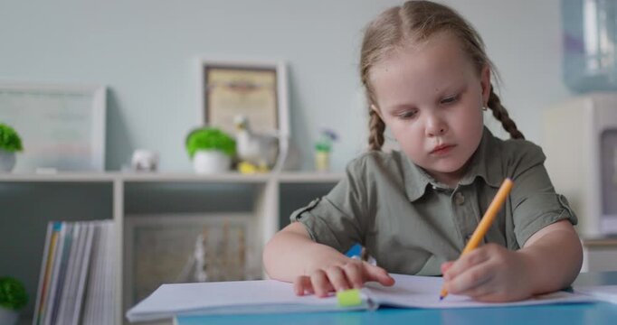 Young schoolgirl with braided hair concentrating on writing in a notebook at a classroom desk, surrounded by shelves, books, and learning supplies