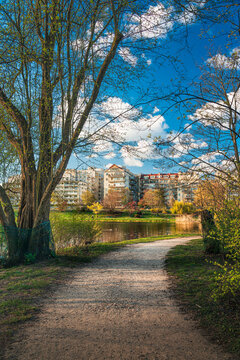 Beautiful spring landscape in Skaryszewski Park in Warsaw, Poland. A gravel pathway leading towards a peaceful lake with modern residential buildings in the background under a blue sky.