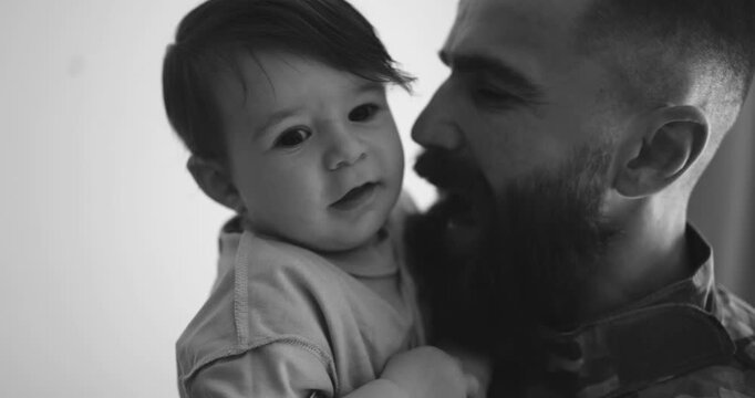 Soldier man in military uniform having tender moment with his little baby son at home - Emotional and happy homecoming concept - UGC content - Black and white editing