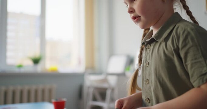 Young girl concentrates on tossing a small ball into red cups during an indoor party game with an adult guiding her at a bright table