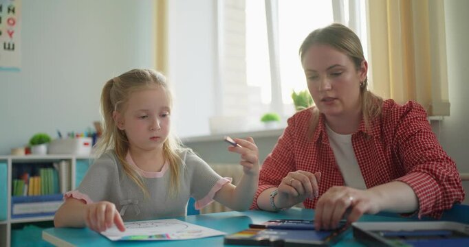 Teacher guiding a young girl during a classroom drawing and writing activity at a blue table in a bright early learning environment