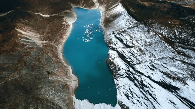 Stunning top-down aerial drone shot of a vibrant turquoise glacial lake nestled between rugged snow-dusted mountain slopes in Nepal's Manaslu region.