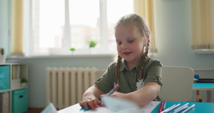 Young girl with braided hair drawing with colored pencils at a classroom desk while another child works nearby in a bright preschool learning space