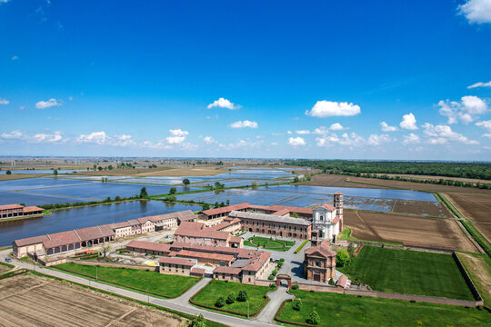 Aerial view of the historic Abbazia di Lucedio complex surrounded by flooded rice fields and green meadows under a clear blue sky in Trino, Piemonte, Italy.