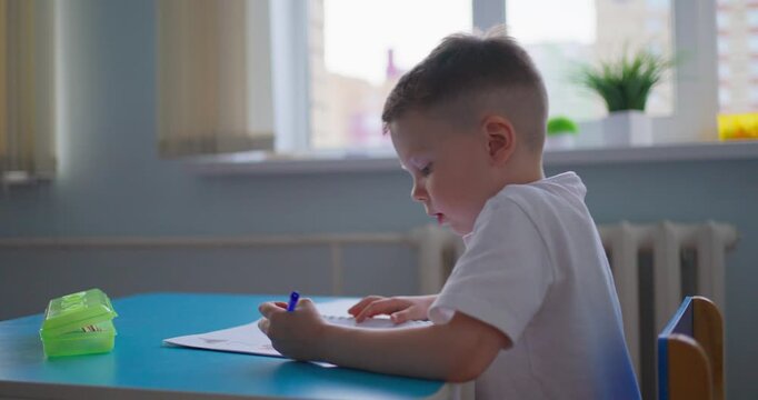 Young boy in a bright classroom writing with a pencil at a blue table, then standing with his notebook during a quiet early learning activity